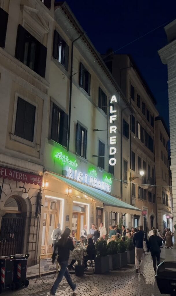 Alfredo alla Scrofa ristorante in Rome at night, with green neon signage, outdoor diners seated at tables, and pedestrians walking the cobblestone street