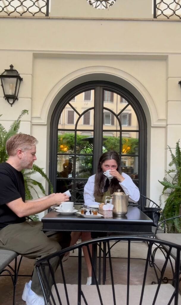 Two people enjoying coffee at an outdoor cafe table in front of an arched window, with ferns and classic Italian architecture in the background
