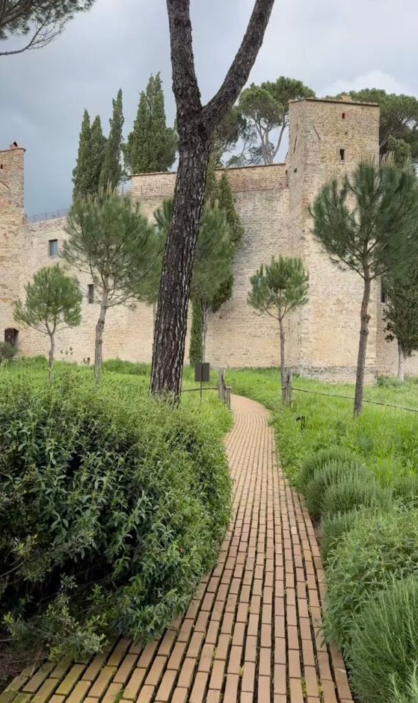 brick garden path leading toward the stone walls of a medieval Italian fortification, flanked by cypress and pine trees under an overcast sky