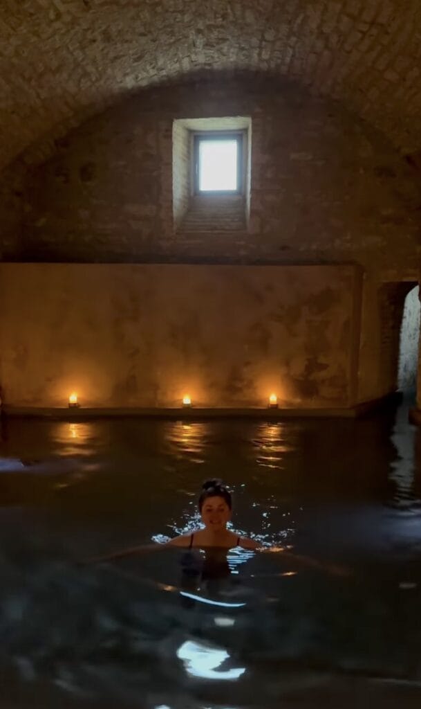 A woman soaking in a candlelit indoor thermal bath with stone vaulted ceiling and small arched window, Italy