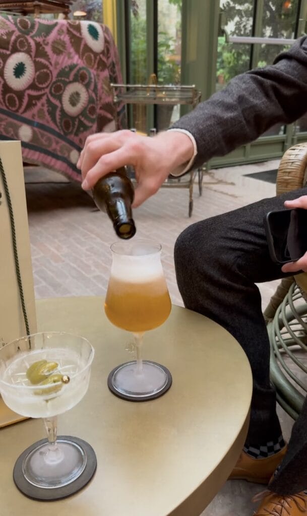 pouring a beer from a bottle into a tulip glass at a cafe table, with a martini and olive coupe glass alongside, rattan chairs and a bright atrium in the background