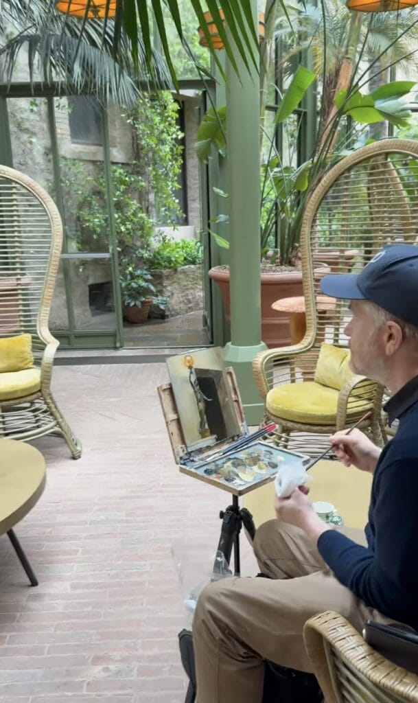 A man painting at a portable easel in a sun-filled conservatory with rattan chairs, yellow cushions, and lush tropical plants