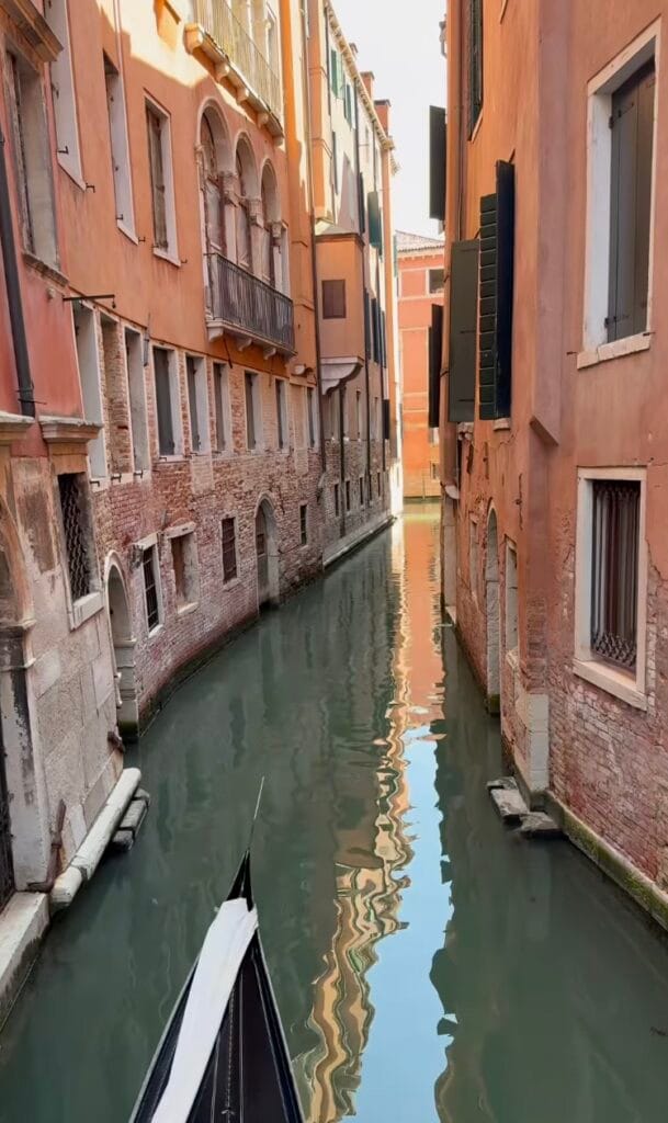 View from a gondola on a narrow Venice canal, lined with terracotta and brick buildings reflecting in the still green water