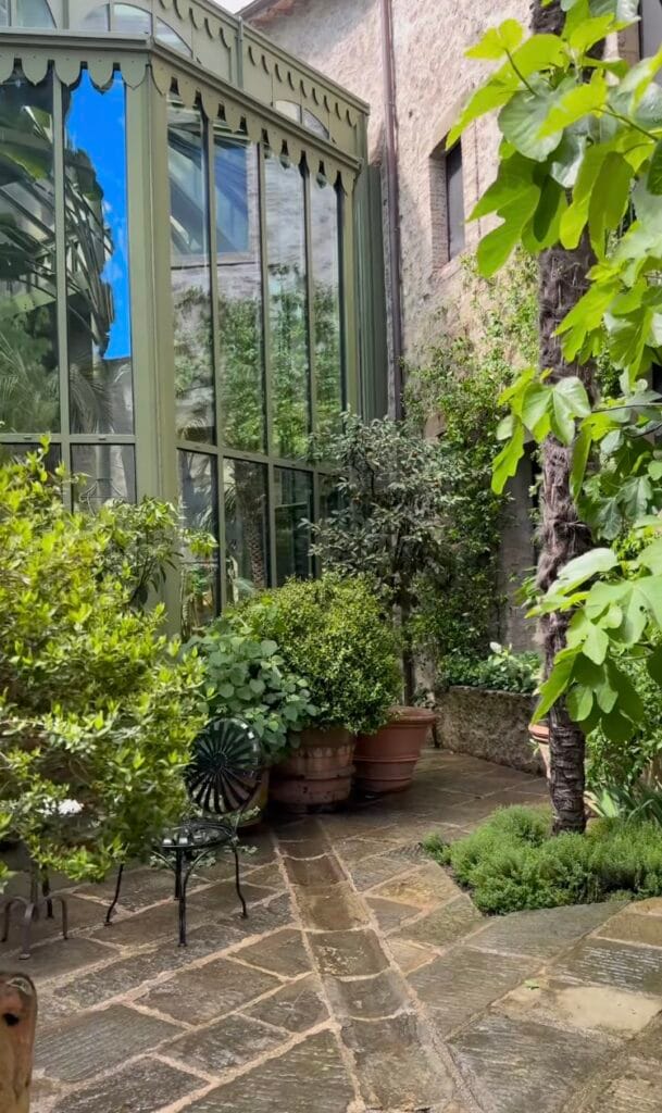 Stone courtyard with a green-framed glass conservatory, potted shrubs in terracotta planters, and a wrought-iron bistro table on a paved path in Italy