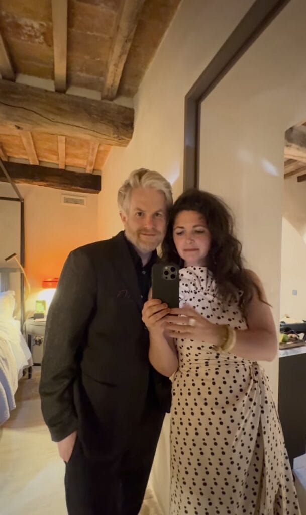 Jordan Ferney and her husband taking a mirror selfie in a rustic Italian room with exposed wooden ceiling beams, she in a cream polka-dot dress, he in a dark suit