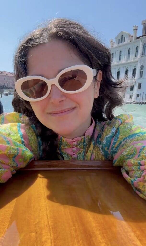 Jordan Ferney resting her arms on a wooden boat in Venice, wearing white oval sunglasses and a colorful paisley jacket, with a canal and grand palazzo in the background