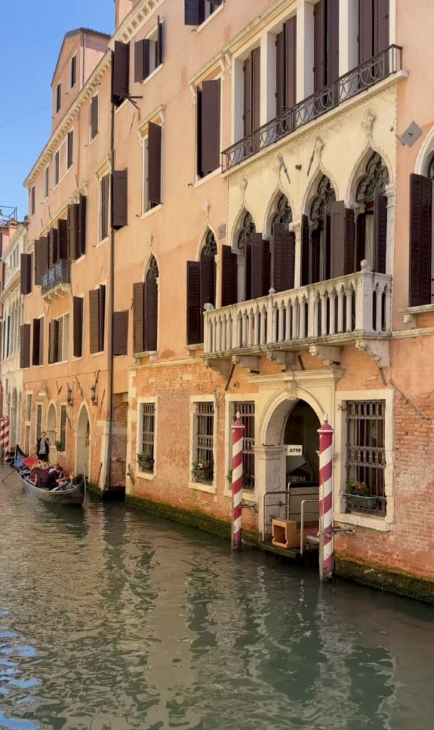 Venice canal with a gondola passing Gothic palazzo facades, dark shutters, an ornate balcony, and an ATM set into an arched water-level doorway