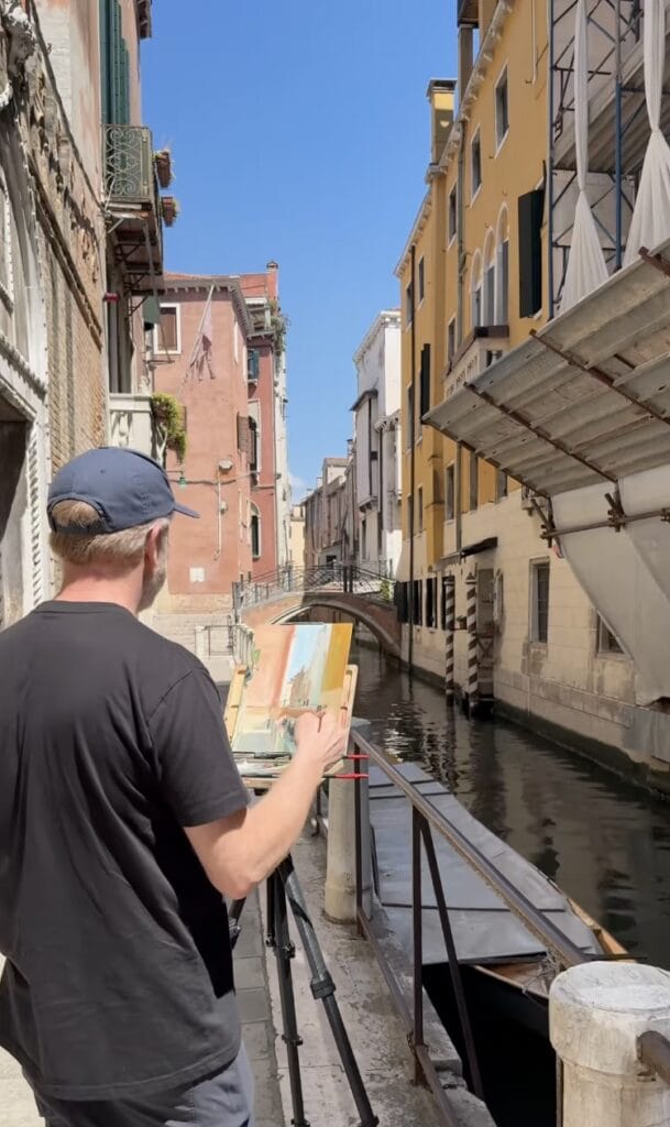 artist painting at an easel beside a Venice canal, with colorful historic buildings and a small bridge in the background