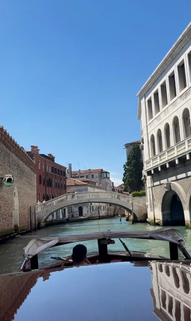 View from a water taxi on a Venice canal, with a stone footbridge ahead and historic buildings lining both sides under a clear blue sky
