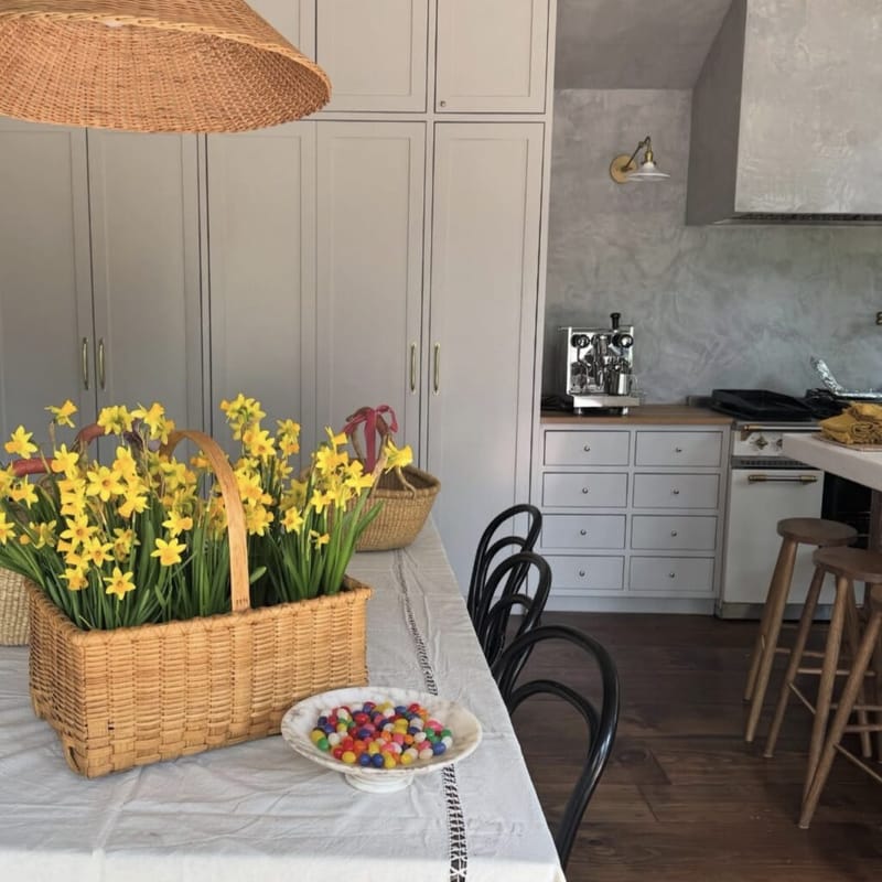 Wicker baskets of yellow daffodils on a linen-covered dining table, with a small bowl of jelly beans, bentwood chairs, and gray kitchen cabinetry with brass hardware in the background