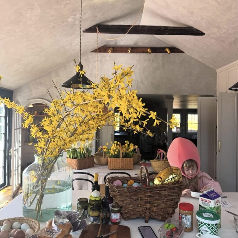 Kitchen counter set up for Easter with a large glass vase of yellow forsythia branches, a wicker basket of colored eggs, and a young child in a pink egg hat seated at the island