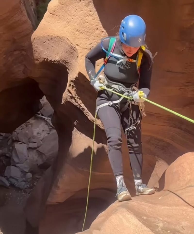 Jordan Ferney rappelling down red sandstone in a canyon at Zion National Park, wearing a blue helmet, wetsuit, harness, and gloves