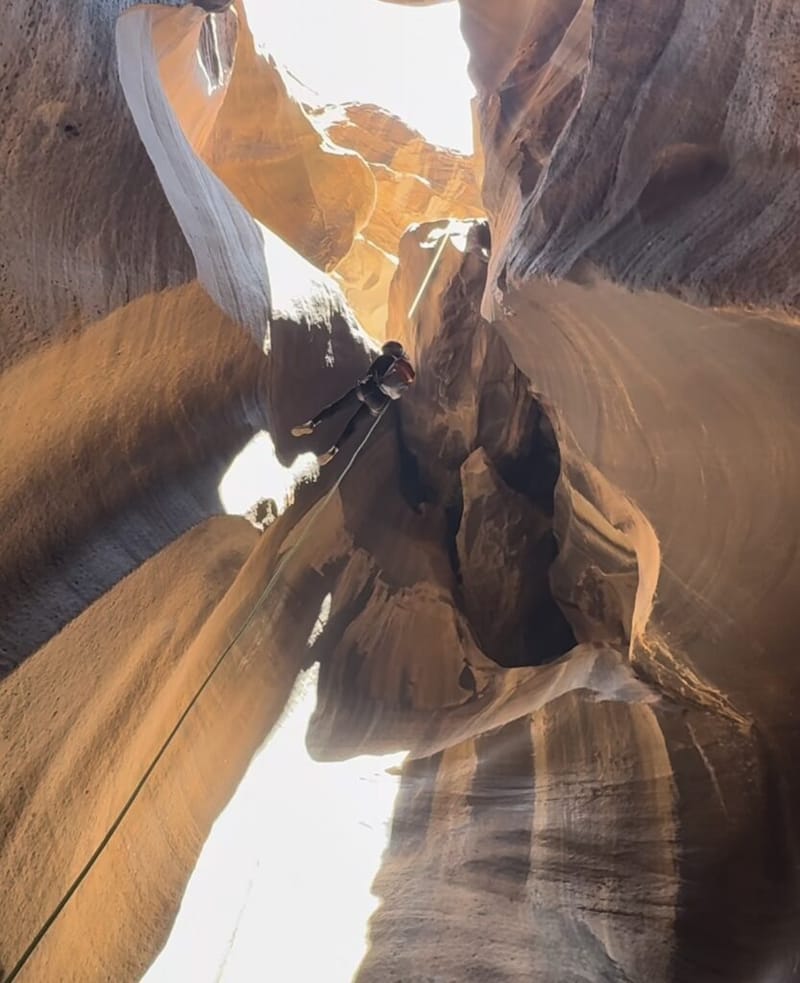 A person rappelling down a narrow sandstone slot canyon at Zion National Park, golden light streaming in from the opening above
