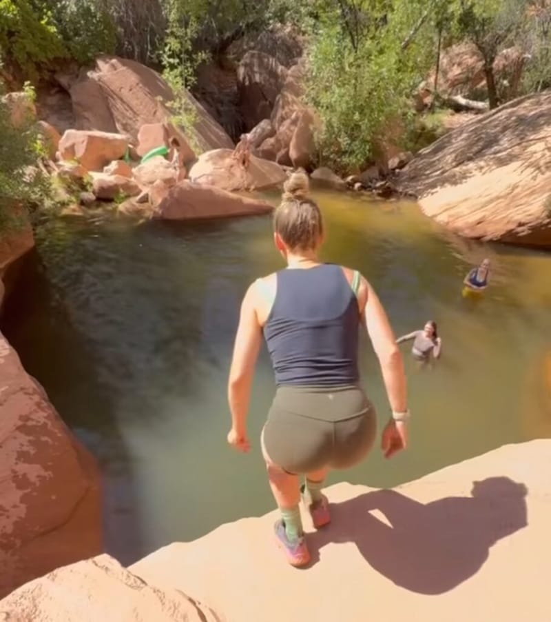 woman on a red sandstone ledge about to jump into a natural swimming hole at Zion National Park, two friends already in the water below
