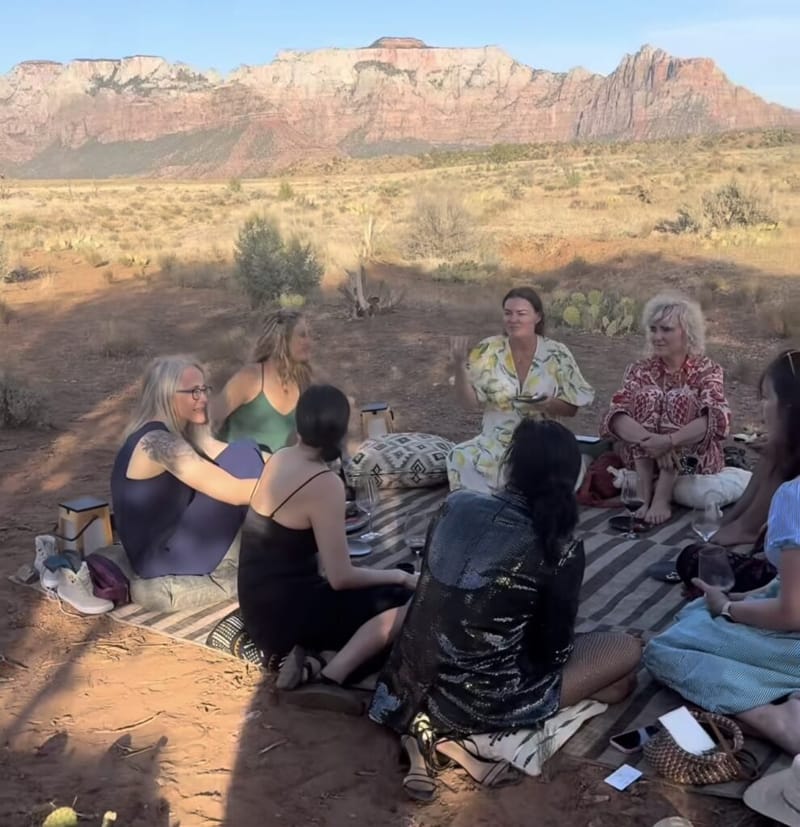 Group of women sitting in a circle on a blanket in a desert clearing, with Zion National Park red rock cliffs in the background