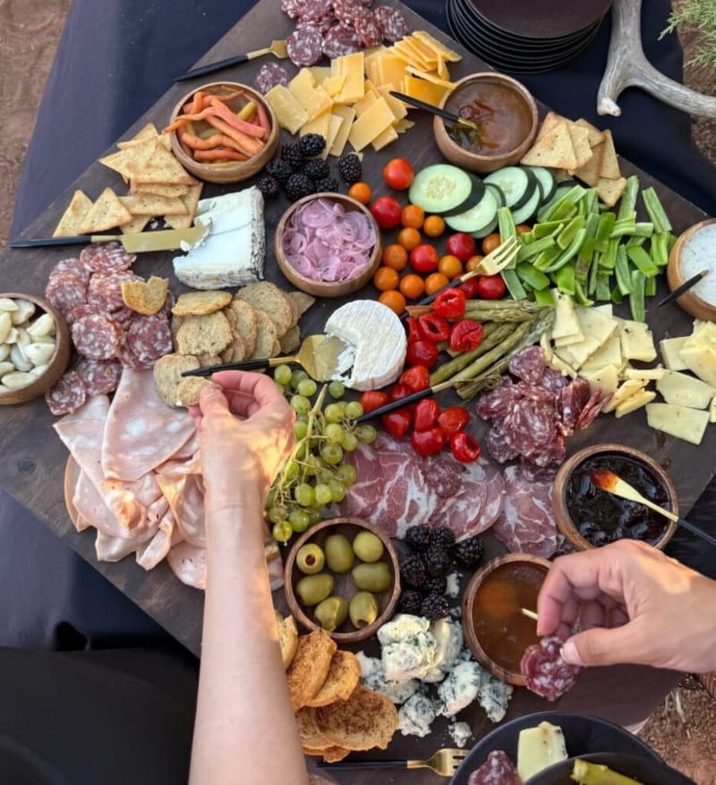 overhead view of a large charcuterie board with cured meats, cheeses, crackers, fresh vegetables, grapes, berries, olives, and dipping sauces, two hands reaching in to graze, set outdoors at Zion National Park