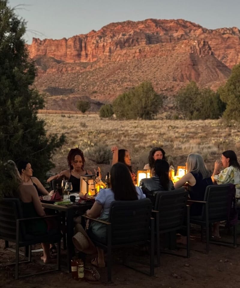 Group of people gathered around a candlelit outdoor dinner table at dusk, red rock canyon cliffs in the background, near Zion National Park
