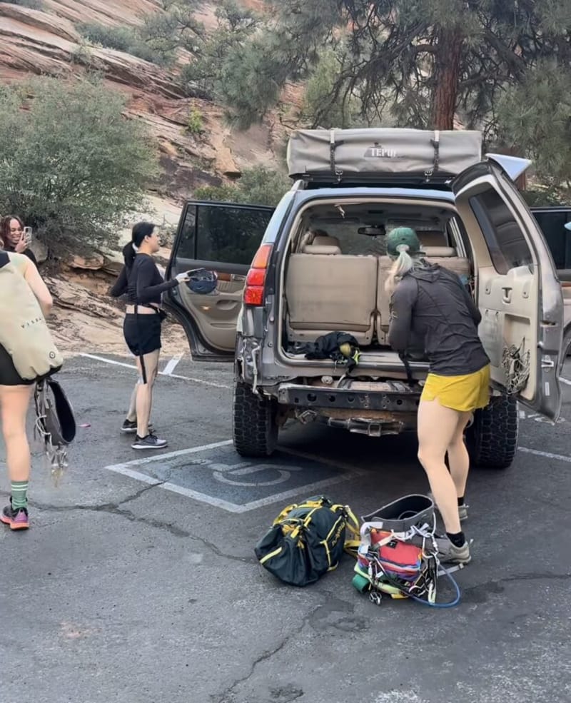 Three people unloading canyoneering gear from an SUV in a Zion National Park parking lot, red sandstone cliffs in the background