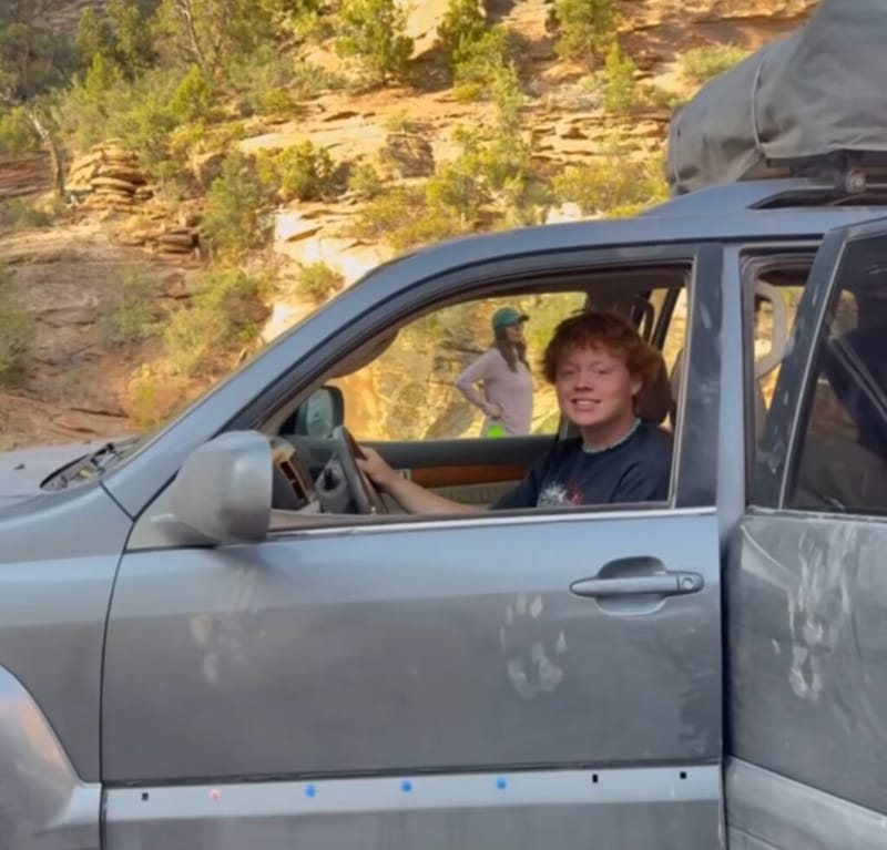A smiling young person with red hair seated in the driver's seat of a silver SUV at Zion National Park, red rock canyon walls and scrub trees visible behind the open door