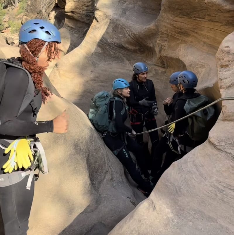 Four canyoneers in helmets, wetsuits, and harnesses gathered in a narrow sandstone slot canyon at Zion National Park