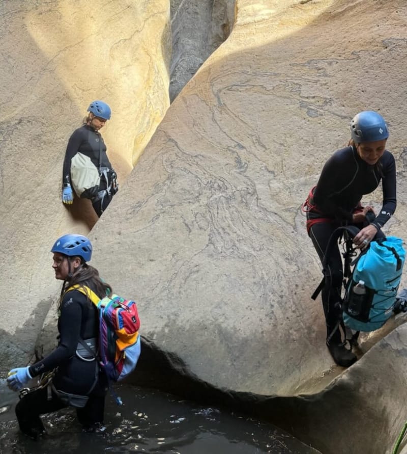three canyoneers in wetsuits and helmets navigating a narrow sandstone slot canyon with water at the base, Zion National Park