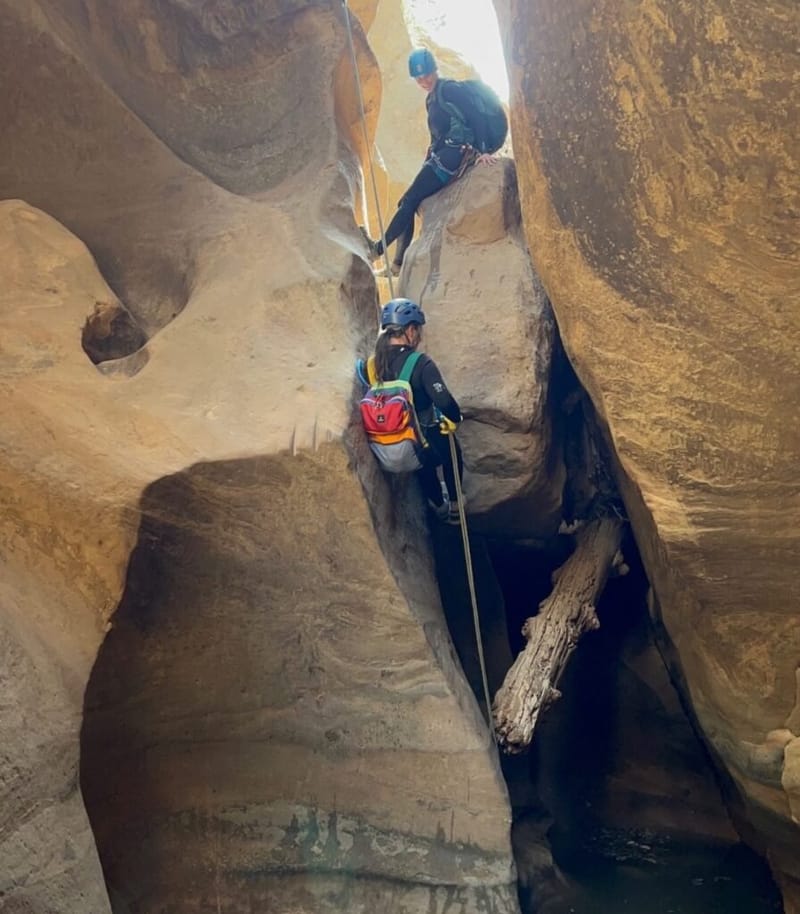 Two canyoneers rappelling through a narrow sandstone slot canyon at Zion National Park, both wearing helmets and harnesses with a rope between them