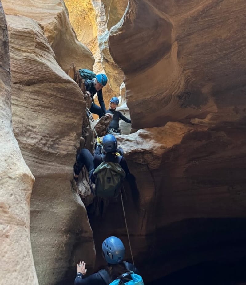 three canyoneers in helmets and wetsuits climbing through a narrow sandstone slot canyon, using a rope to navigate a large rock obstacle