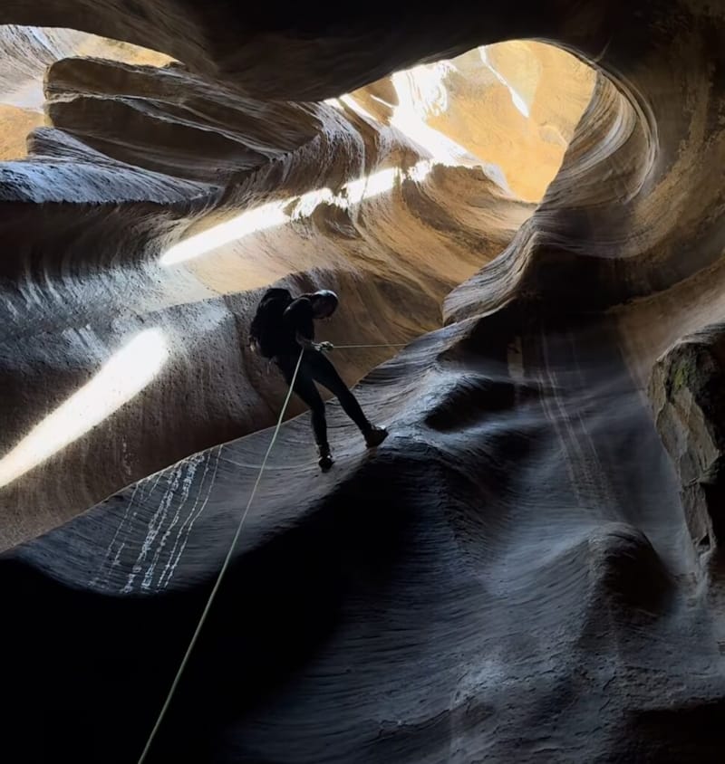 A canyoneer rappelling into a narrow slot canyon in Zion National Park, silhouetted against swirling sandstone walls with a beam of sunlight streaming from the opening above
