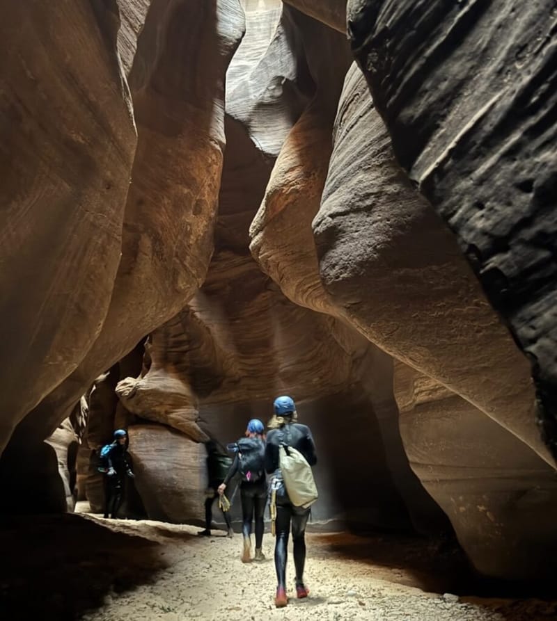 three hikers in wetsuits and blue helmets walking through a narrow slot canyon at Zion National Park, tall smooth sandstone walls rising on both sides