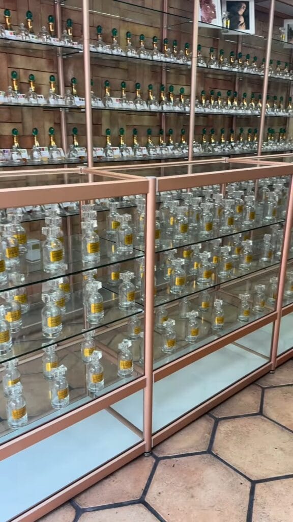 Wall-to-wall glass shelving units filled with hundreds of small mezcal bottles in a Mexico City shop, rose-gold metal frames, terracotta tile floor