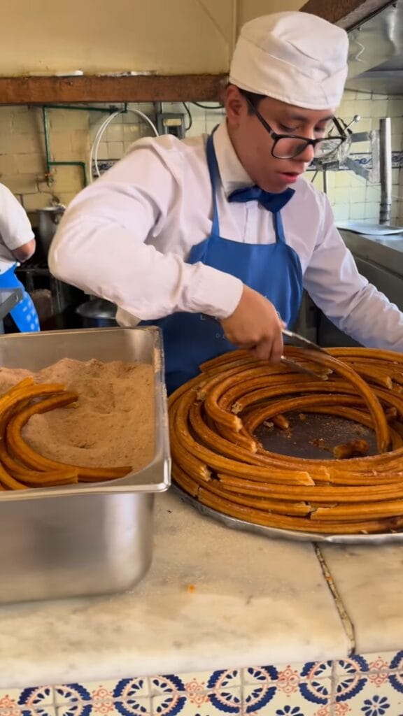 A kitchen worker in a white uniform and blue apron cutting a large coiled spiral of freshly fried churros on a counter, with a tray of cinnamon sugar beside it