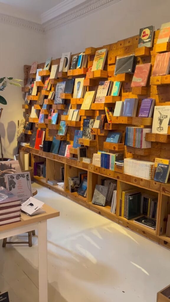 A wood block wall display covered in face-out books inside a bookshop, with a lower shelf row and a small side table stacked with books