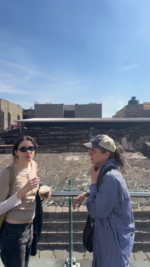 Two women chatting on a terrace overlooking the Templo Mayor ruins in Mexico City, one holding a coffee cup