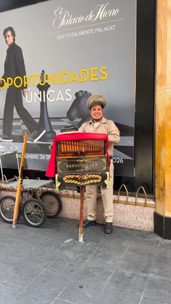 Street musician in tan uniform and cap posing with a hand-cranked barrel organ in front of an El Palacio de Hierro storefront in Mexico City