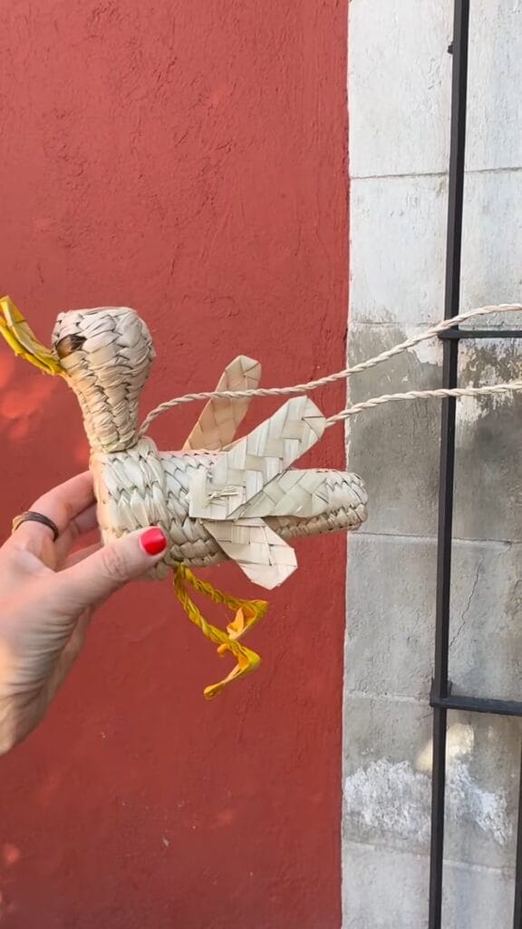 Hand holding a woven palm-leaf bird figurine in front of a red stucco wall in Mexico City