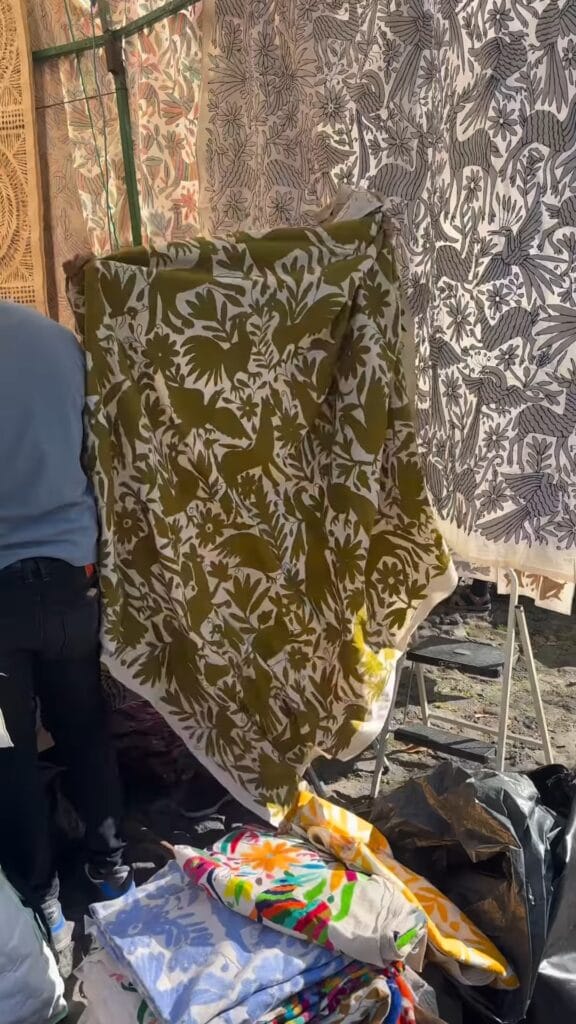 Olive green and cream otomi-patterned textile held up at an outdoor market stall in Mexico City, with additional folk-art printed fabrics hanging in the background