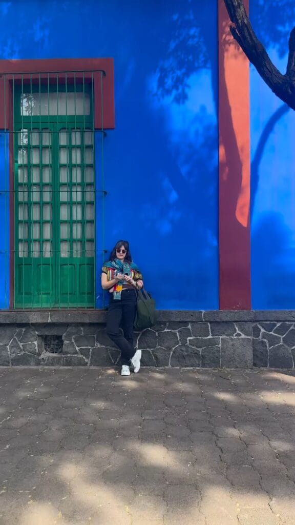 Jordan Ferney leaning against the cobalt blue exterior wall of Casa Azul, the Frida Kahlo Museum in Mexico City, with a green barred door and red trim behind her