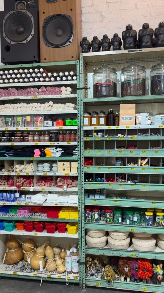 Densely stocked market shelves in Mexico City holding honey jars, piloncillo, dried goods in glass jars, colorful containers, gourds, and clay figurines, with a large wooden speaker in the corner
