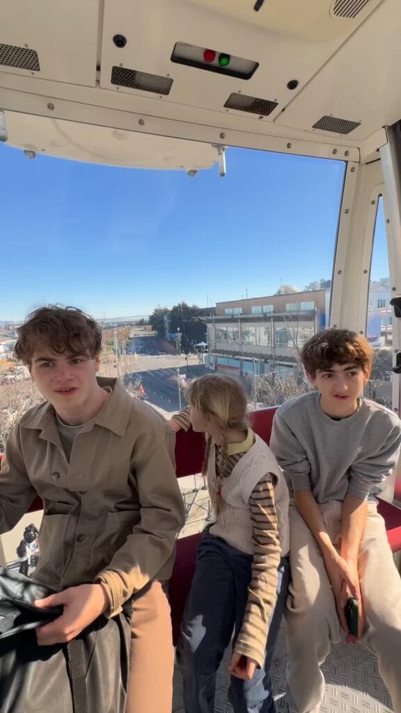 Three kids inside a Ferris wheel gondola, city skyline and blue sky visible through the glass behind them