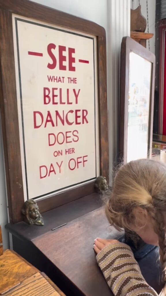 Vintage framed sign reading "See What the Belly Dancer Does on Her Day Off" in a San Francisco antique shop, with a young girl leaning over the display ledge and small bronze busts on the shelf