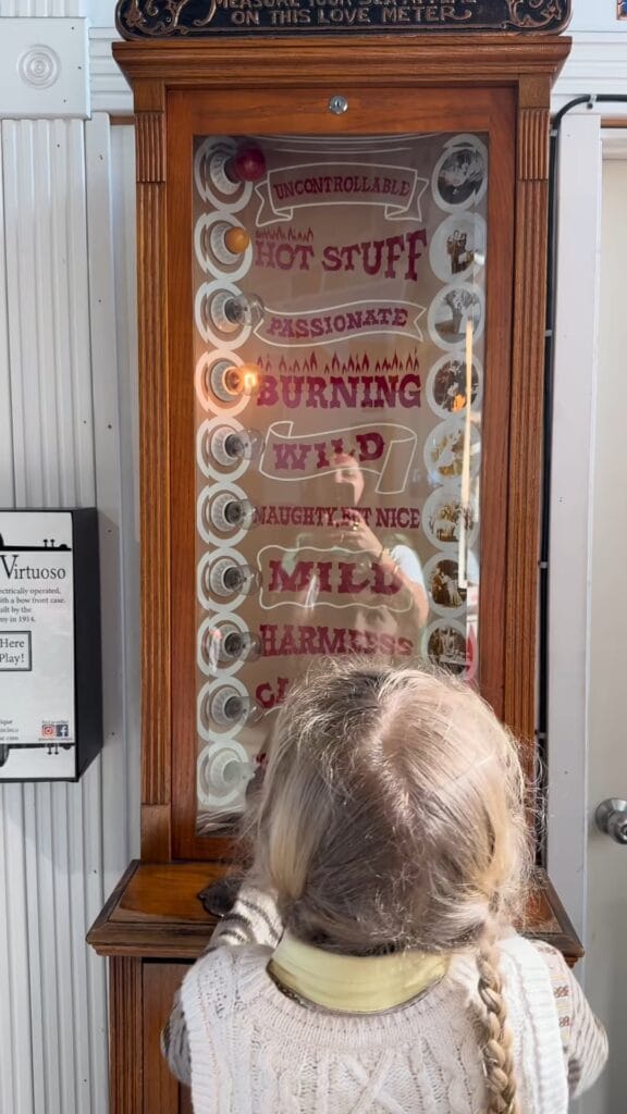 A child with a blonde braid stands facing a vintage Love Meter arcade machine, with temperature labels ranging from Cold to Uncontrollable on the glass panel