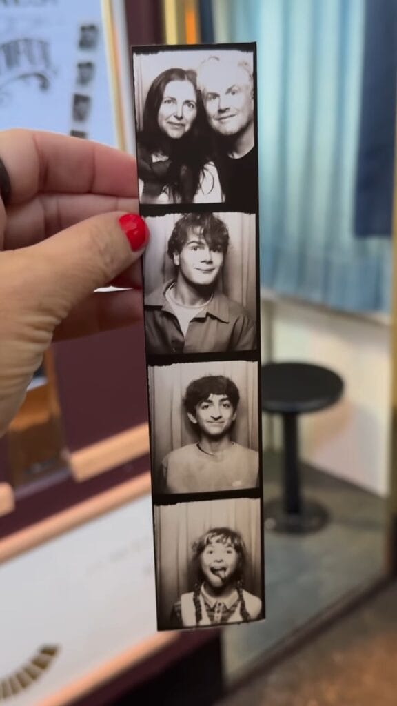 hand holding a black-and-white photo booth strip with four frames, a couple at top, two teenage portraits in the middle, and a young girl sticking out her tongue at the bottom