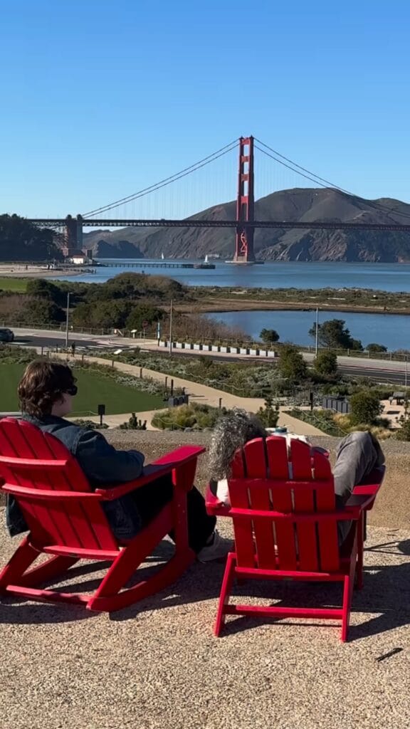 Two people sitting in red Adirondack chairs with a dog, overlooking the Golden Gate Bridge and San Francisco Bay from the Presidio