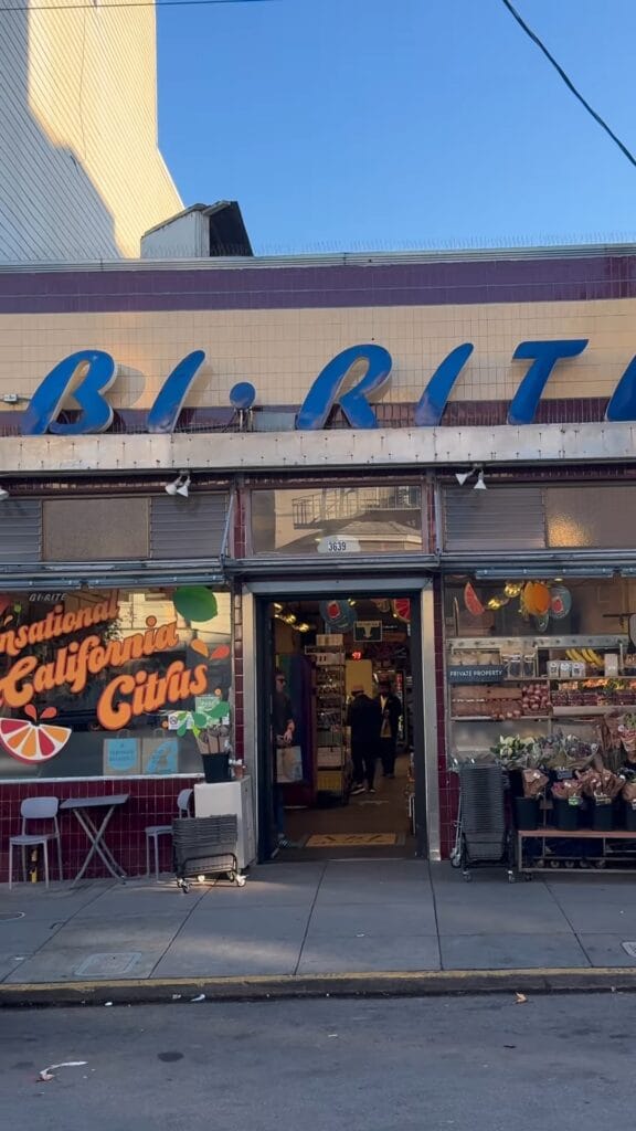 Bi-Rite Market storefront in San Francisco's Mission District, with blue neon sign, a California Citrus window display, and fresh flowers outside