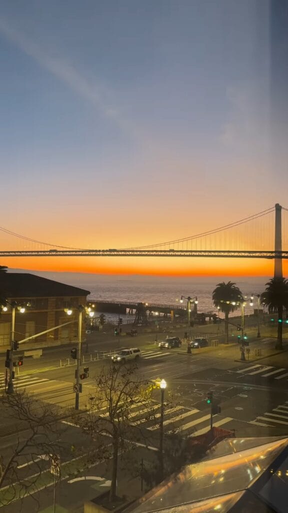 Sunset view of the San Francisco Bay Bridge from the Embarcadero, with orange sky, palm trees, and street traffic below