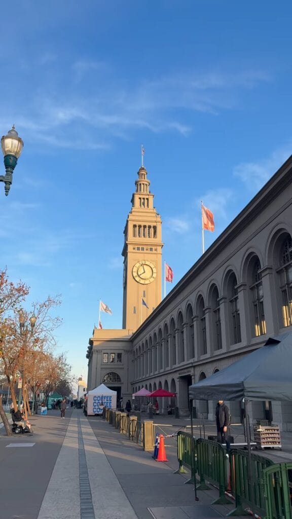 San Francisco Ferry Building clock tower and Embarcadero promenade on a clear day, with farmers market tents along the walkway
