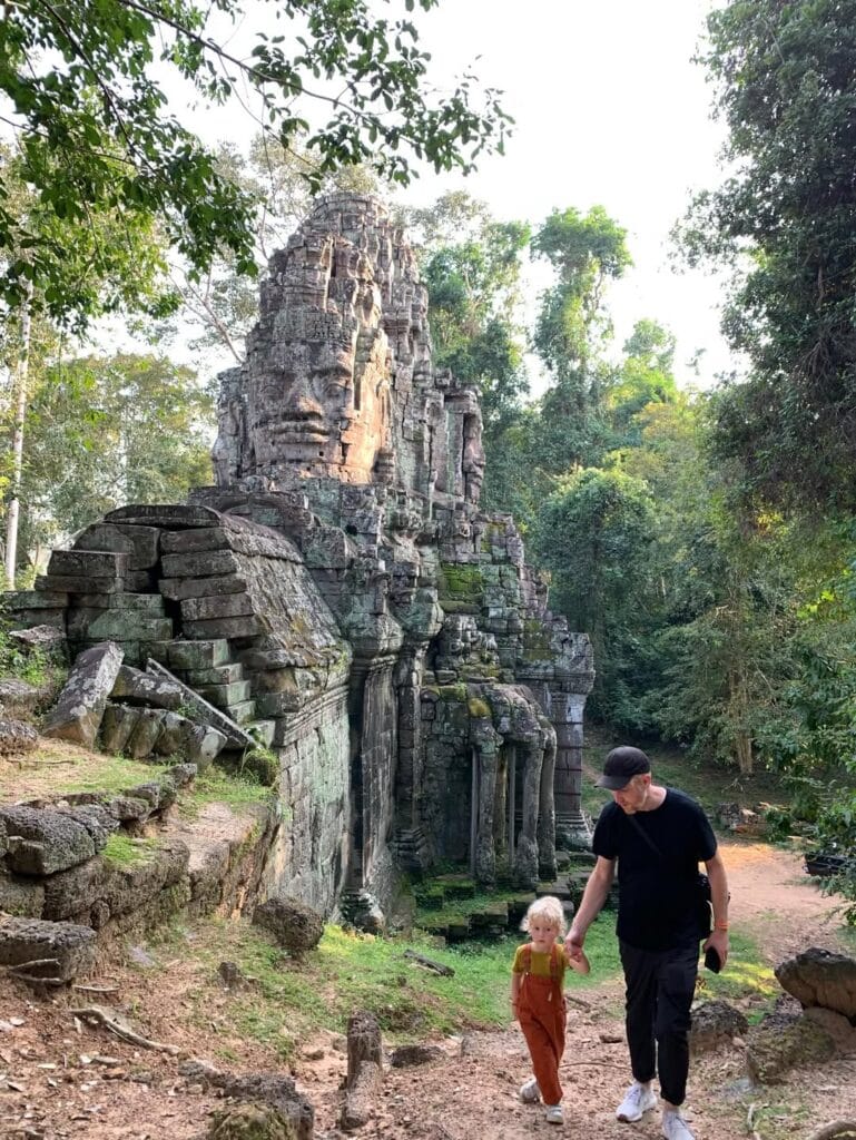 A man and young child walking hand in hand through a carved stone temple gate at the Angkor complex in Cambodia, with large stone faces carved into the tower above them