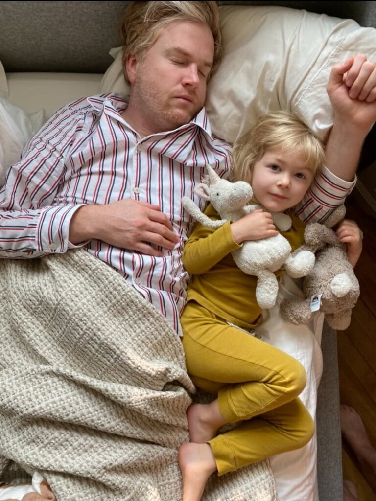 Jordan Ferney napping in bed with a toddler in mustard pajamas clutching two stuffed animals