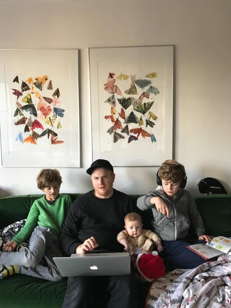 Paul Ferney on a green sofa with three kids, a MacBook on his lap, two framed butterfly prints on the wall behind them