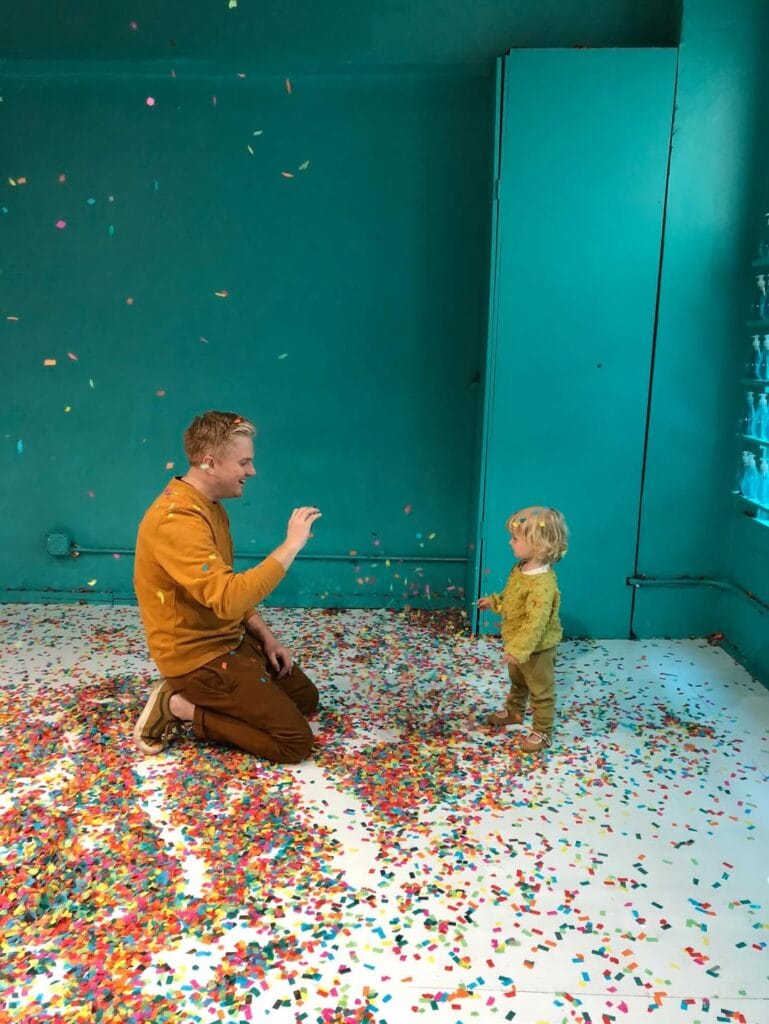 A man kneeling on a confetti-covered floor laughing and playing with a toddler in a teal-walled room